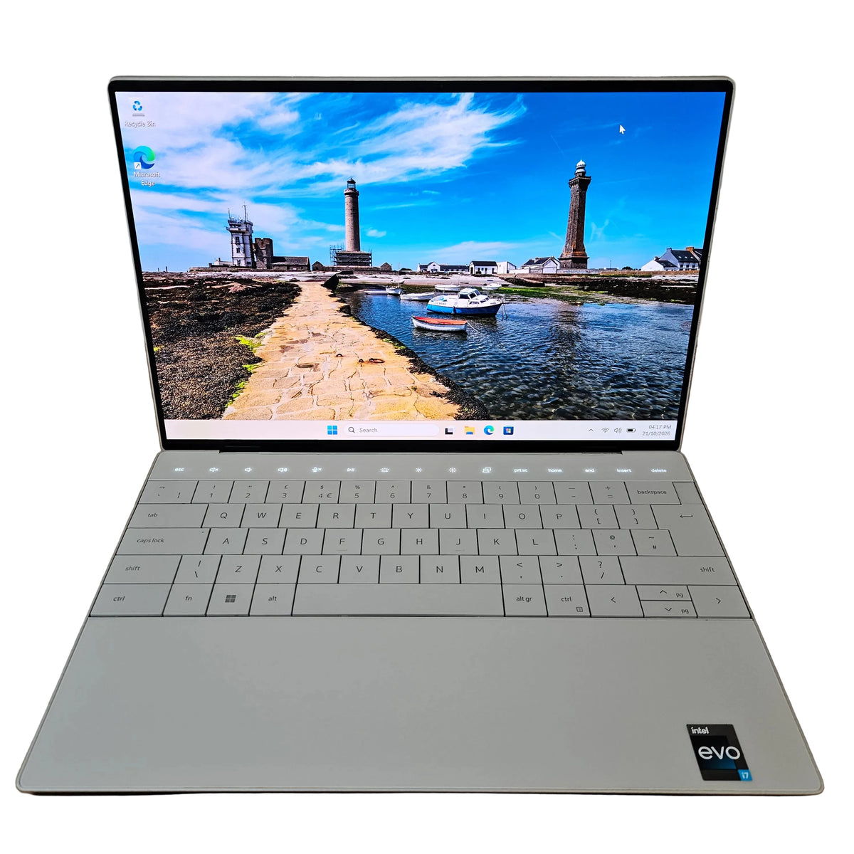 A silver dell xps 9320 EVO laptop is open and photographed from above, resting on a brown cardboard box against a light gray wall. The screen is on and displays a harbor scene with lighthouses.