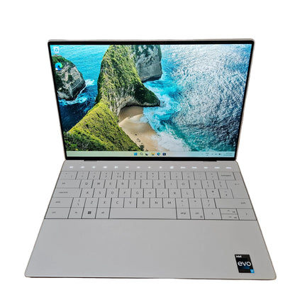 A silver dell xps 9320 laptop is open and placed on a cardboard box on a carpeted floor against a white wall. The screen displays a scenic wallpaper of a coastal cove.